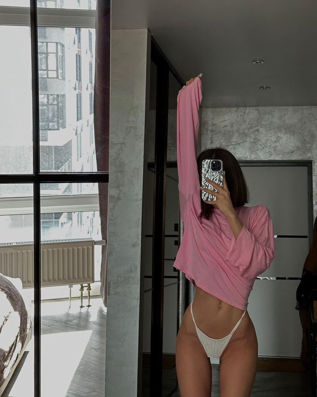 A woman stands by large windows in a cropped pink top and white bottoms, taking a mirror selfie with a raised arm.