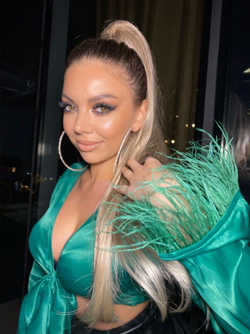 A woman with a blonde ponytail wears a teal satin top with feathered shoulders, hand near hair against a dark background.