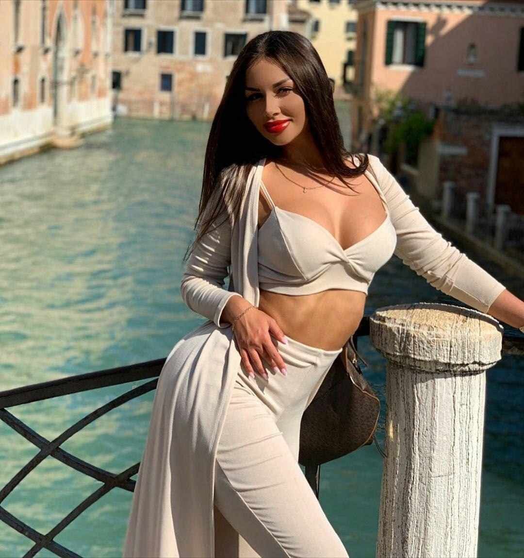 Olivia stands on a bridge over a canal, wearing a light cropped top, high-waisted pants, and an open long cardigan.
