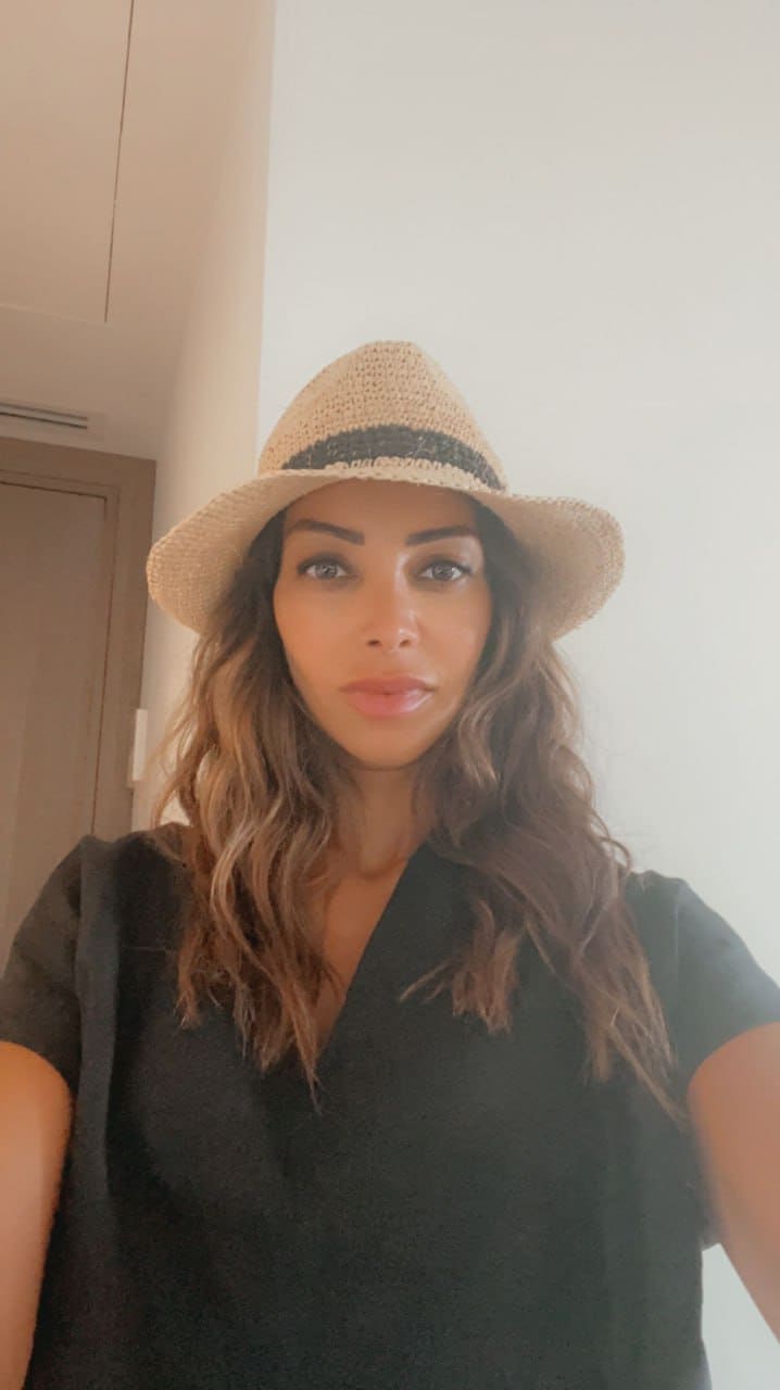 Woman in straw fedora and black short-sleeved top stands indoors near a white wall and wooden door.