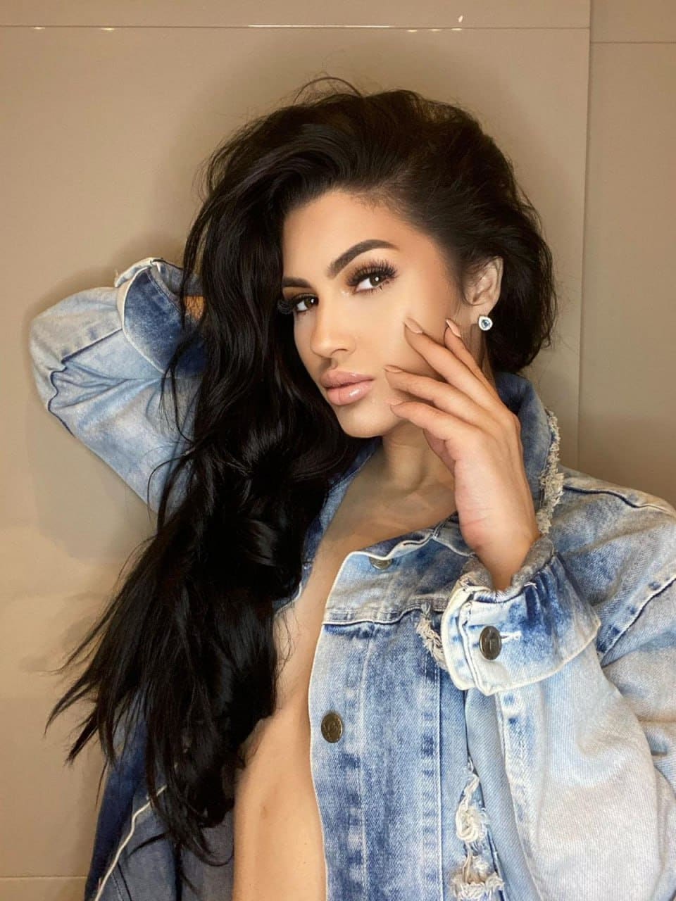 Olivia poses against a beige wall in an open distressed denim jacket, one hand raised to her cheek and the other behind her head.