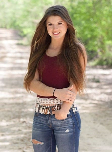 Béatrice stands on a dirt path in a maroon tank top, lace belt, and distressed jeans, with one hand on her waist.