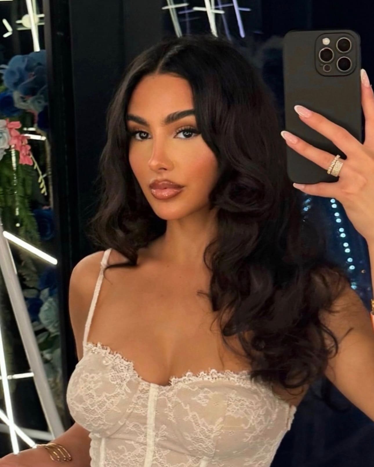 A mirror selfie shows a woman in a white lace bodysuit holding a phone, against a dark background with string lights.