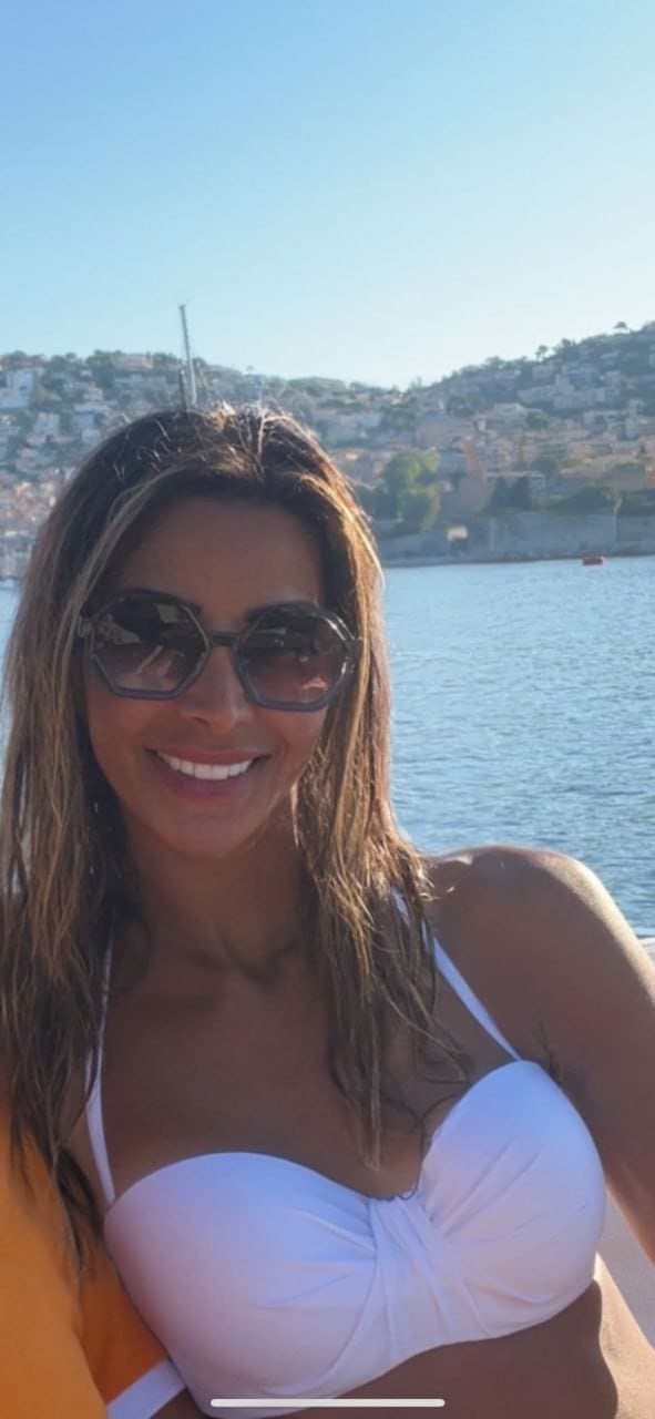 Woman in white bikini top and dark sunglasses smiles outdoors, with water and a hillside town behind her.