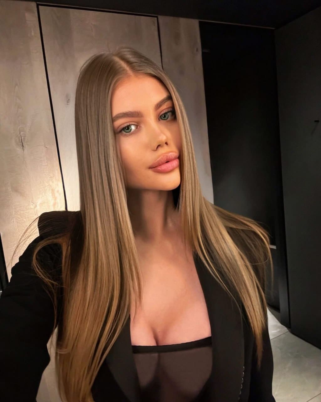 Mila takes a selfie in a black blazer over a sheer top, standing against dark paneled walls.