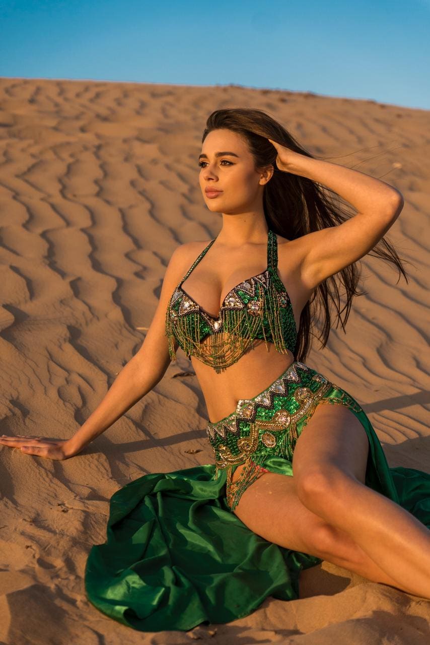 A woman sits on a sand dune under a blue sky, wearing a green and gold beaded two-piece costume with a flowing skirt.