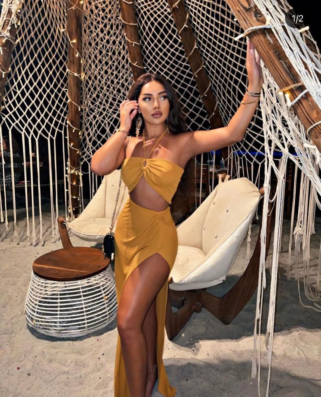 A woman stands on sand in a mustard-yellow cut-out dress with a thigh slit, touching a rope structure, with white chairs and a table behind.