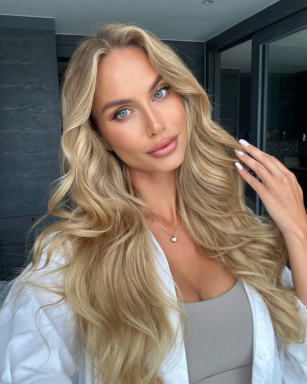 Lily poses indoors with long wavy blonde hair, wearing a white open shirt over a beige ribbed top and a thin necklace, hand raised near her hair.