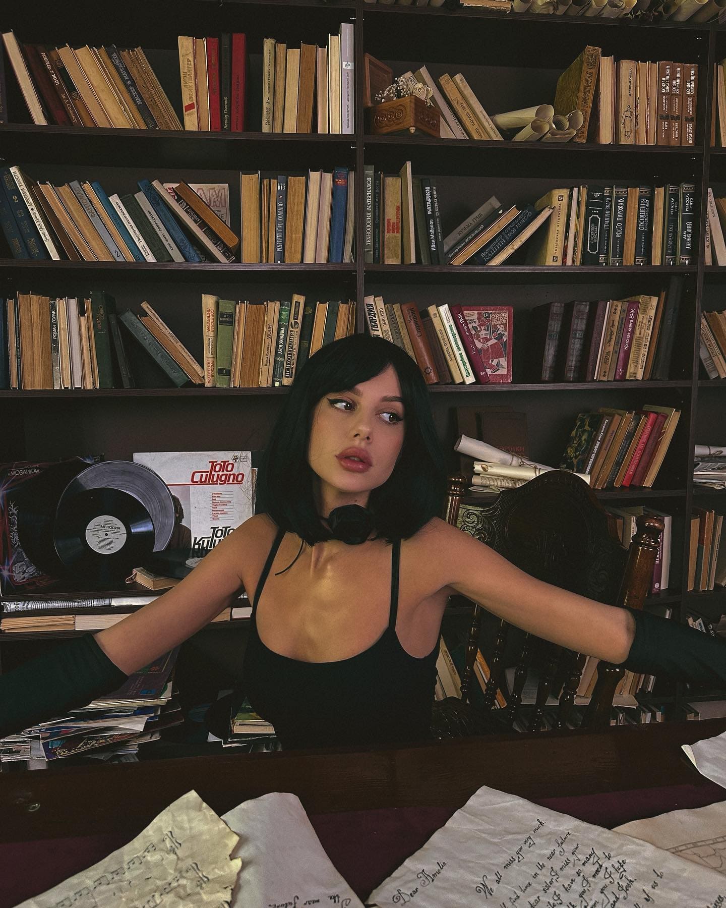 A woman sits at a desk before bookshelves in a black spaghetti-strap top and long black gloves, with a vinyl record and papers on the surface.