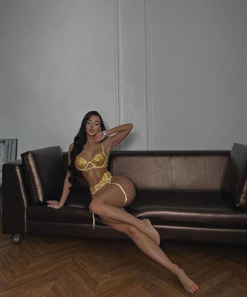 A woman sits on a brown leather sofa in a gold lace two-piece set, one hand behind her head and legs extended on the wooden floor.