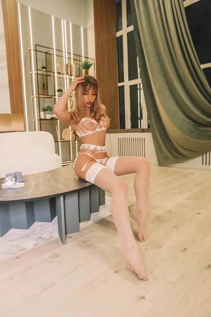A woman sits on a low table in white lace lingerie with sheer panels and stockings, raising one hand to her head in a modern room.