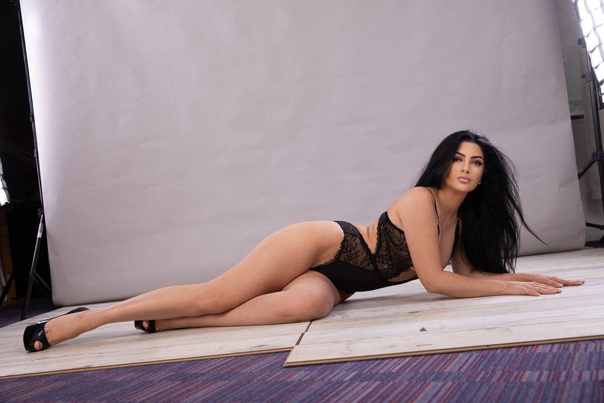 A woman lies on her side on a wooden studio floor in a black lace bodysuit and heels, with a white backdrop and lighting equipment behind her.