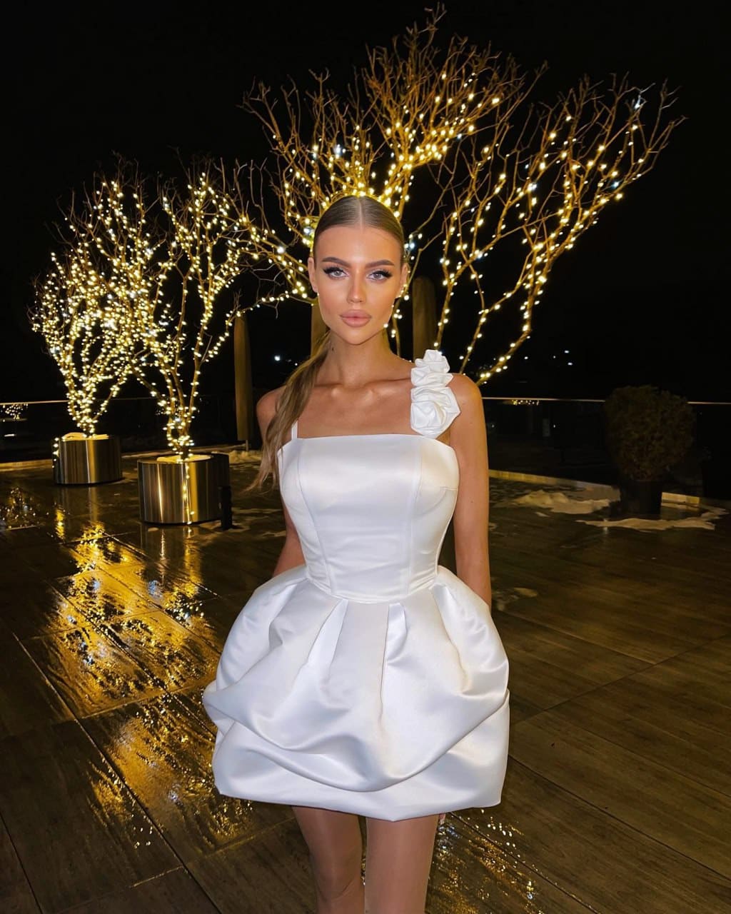 A blonde woman in a white satin mini dress stands on a wet deck at night, illuminated trees behind her.