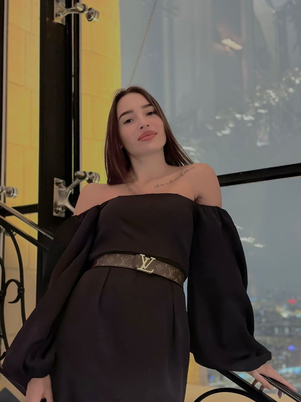 Zora stands on an indoor staircase in a black off-the-shoulder dress with wide sleeves and a patterned belt, with a cityscape visible through a large window behind her.