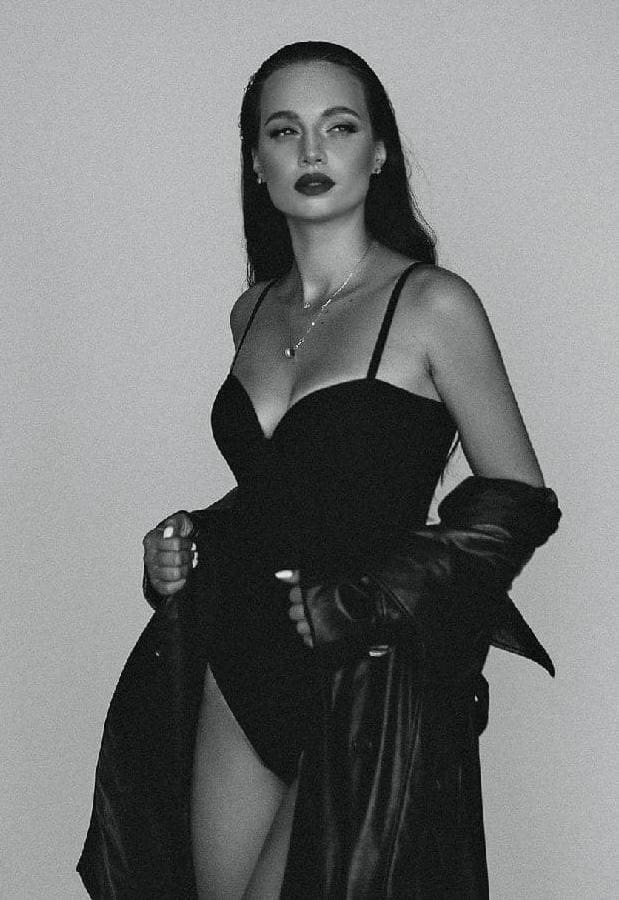 A woman stands in a studio wearing a black slip dress with a deep V-neck and high slit, with a dark jacket draped off her shoulders.