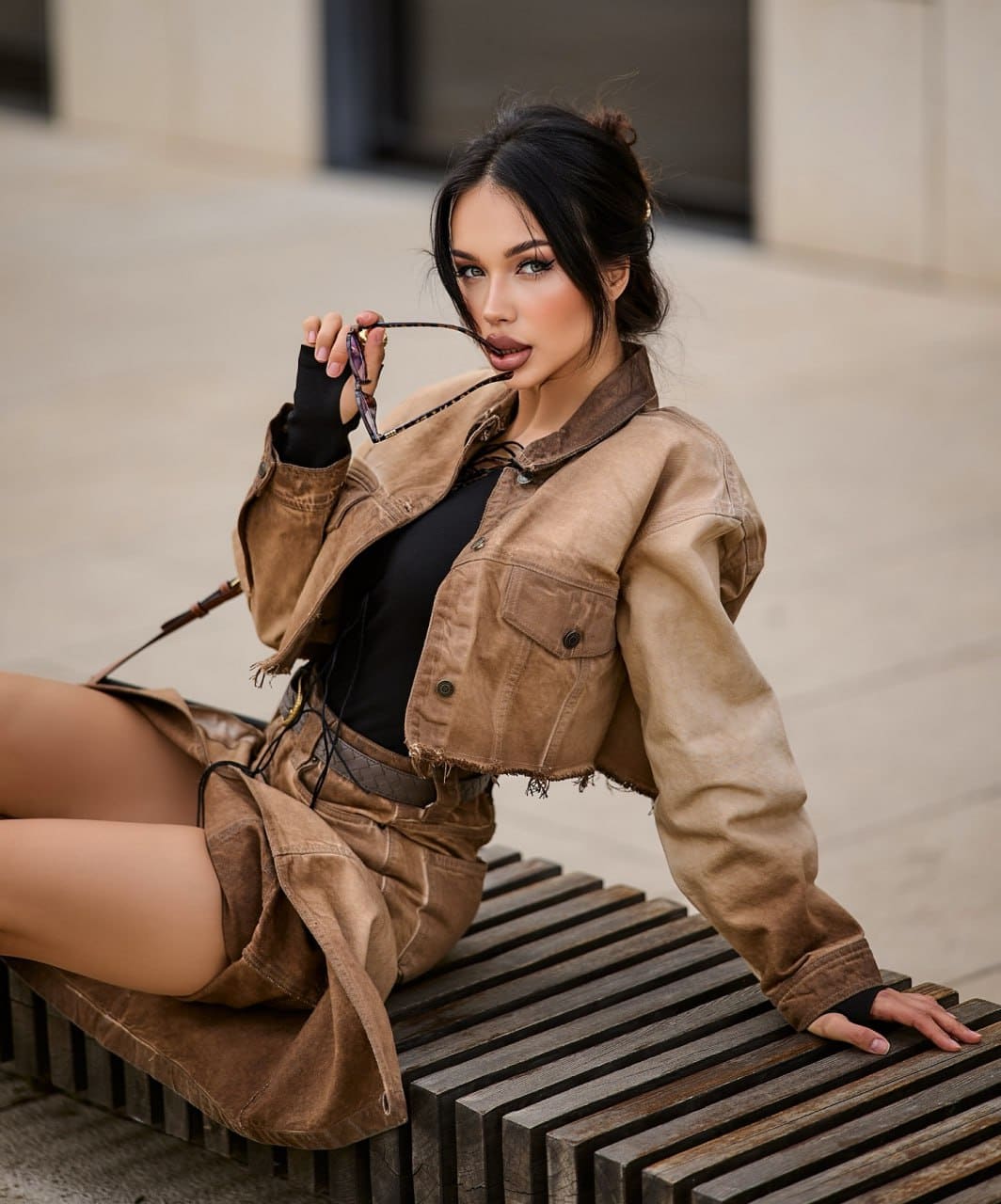 A woman in a tan cropped jacket and skirt sits on a wooden bench outdoors, holding sunglasses and looking at the camera.