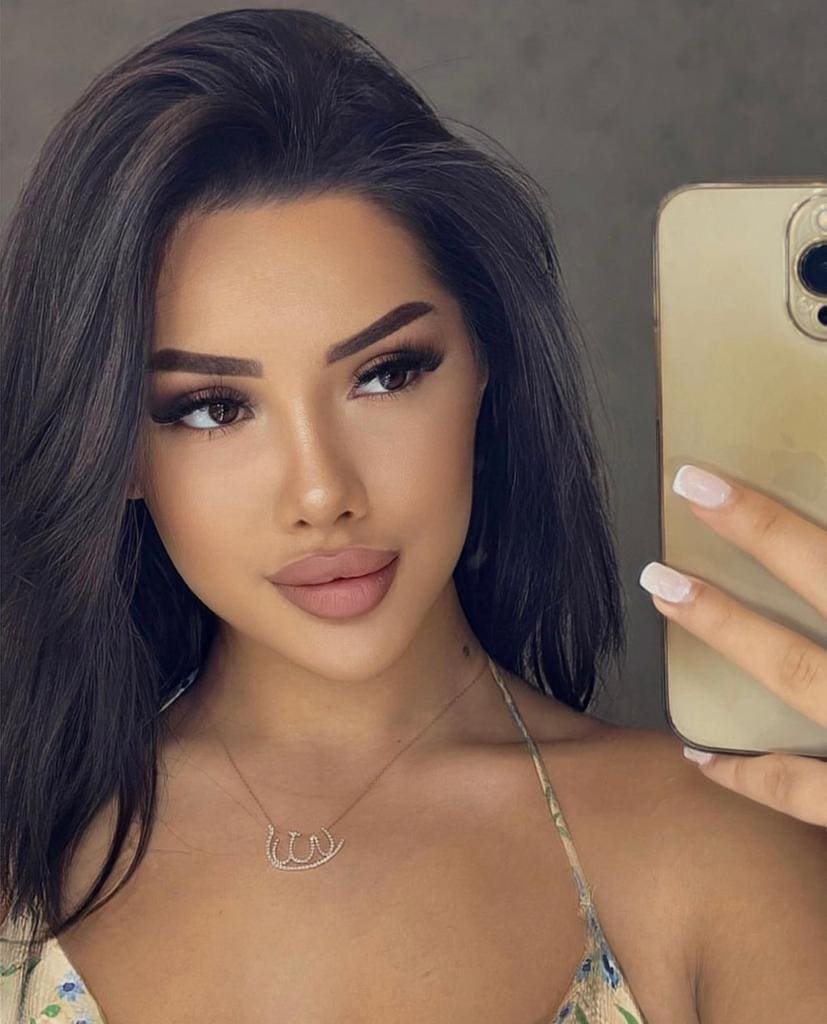 Leila takes a mirror selfie with a gold phone, wearing a thin-strapped floral top and necklace against a gray wall.