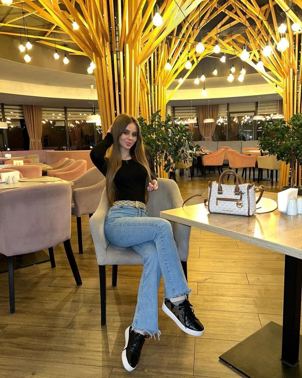 A woman in a black long-sleeve top and jeans sits on a grey chair in a restaurant, touching her hair near a handbag.