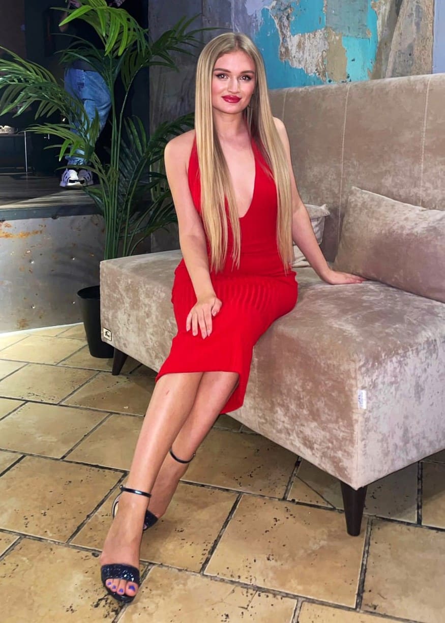 A woman in a red sleeveless midi dress and black heels sits on a beige velvet sofa, with a plant and tiled floor behind her.